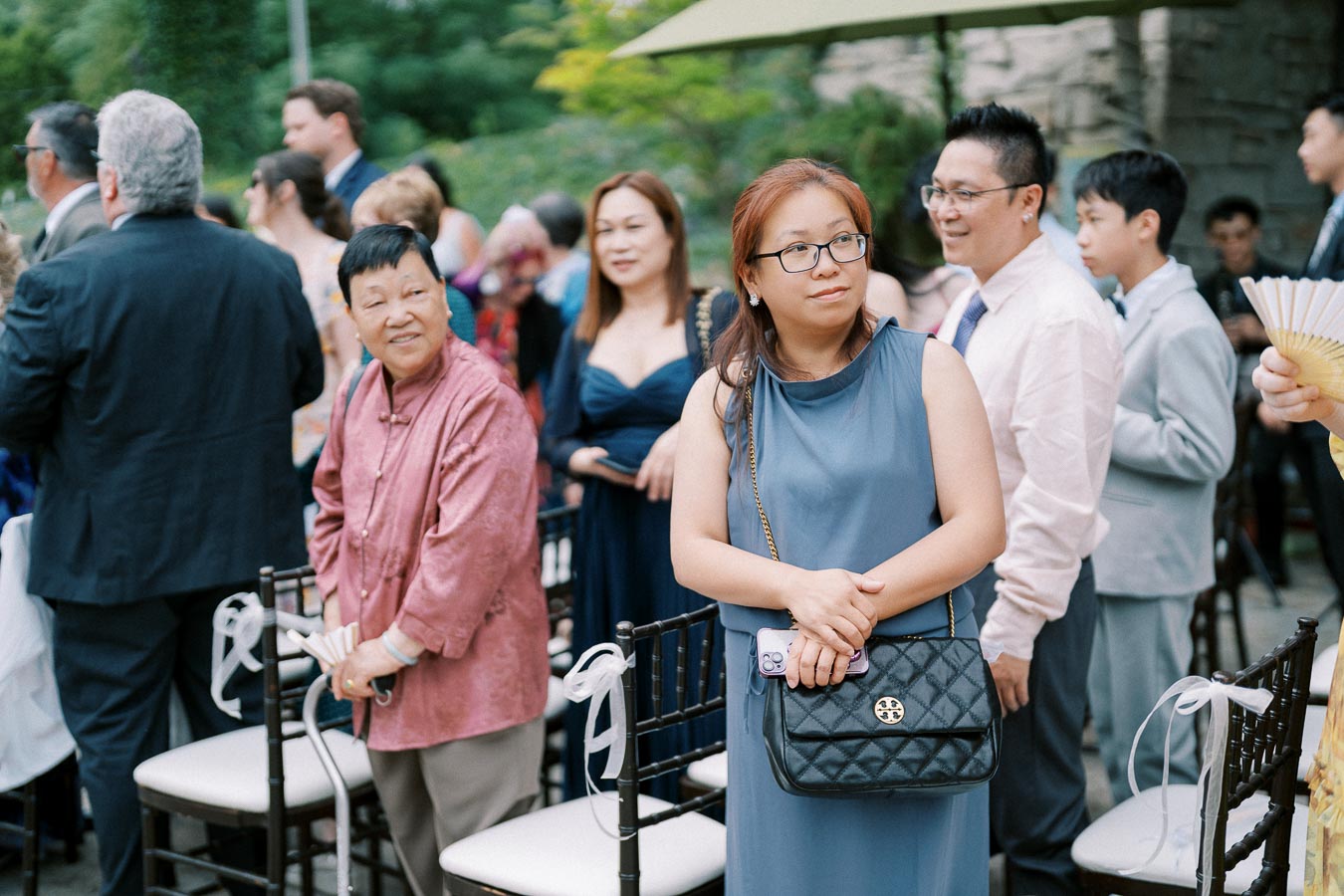 A group of people attending an outdoor event, dressed in formal attire, some wearing suits and dresses, with one woman in focus holding a black purse and smartphone.