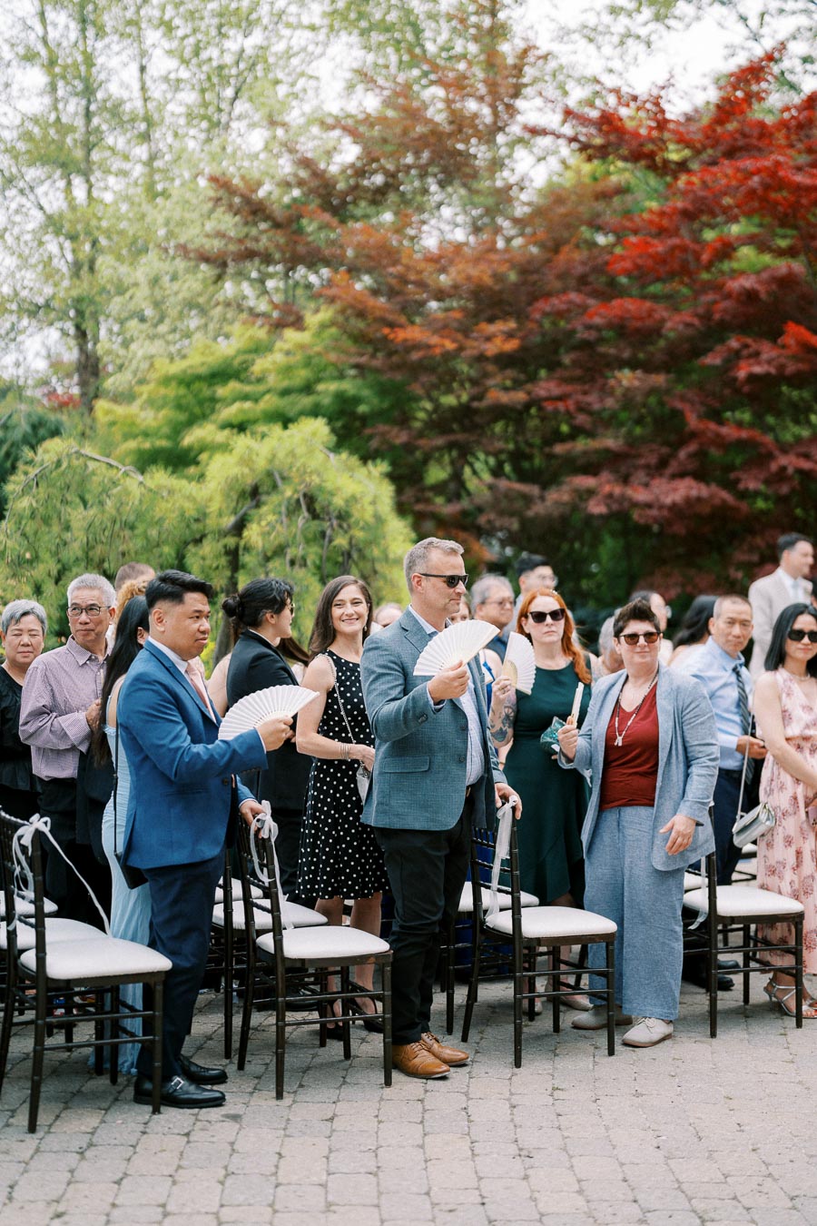 A group of well-dressed wedding guests standing outdoors, holding handheld fans, during a daytime ceremony. The setting features lush greenery and colorful foliage, reflecting a festive and elegant garden atmosphere.
