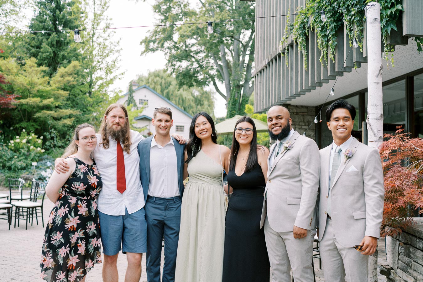 Group of seven people posing together at an outdoor event, dressed in formal and semi-formal attire, with greenery and a building in the background.