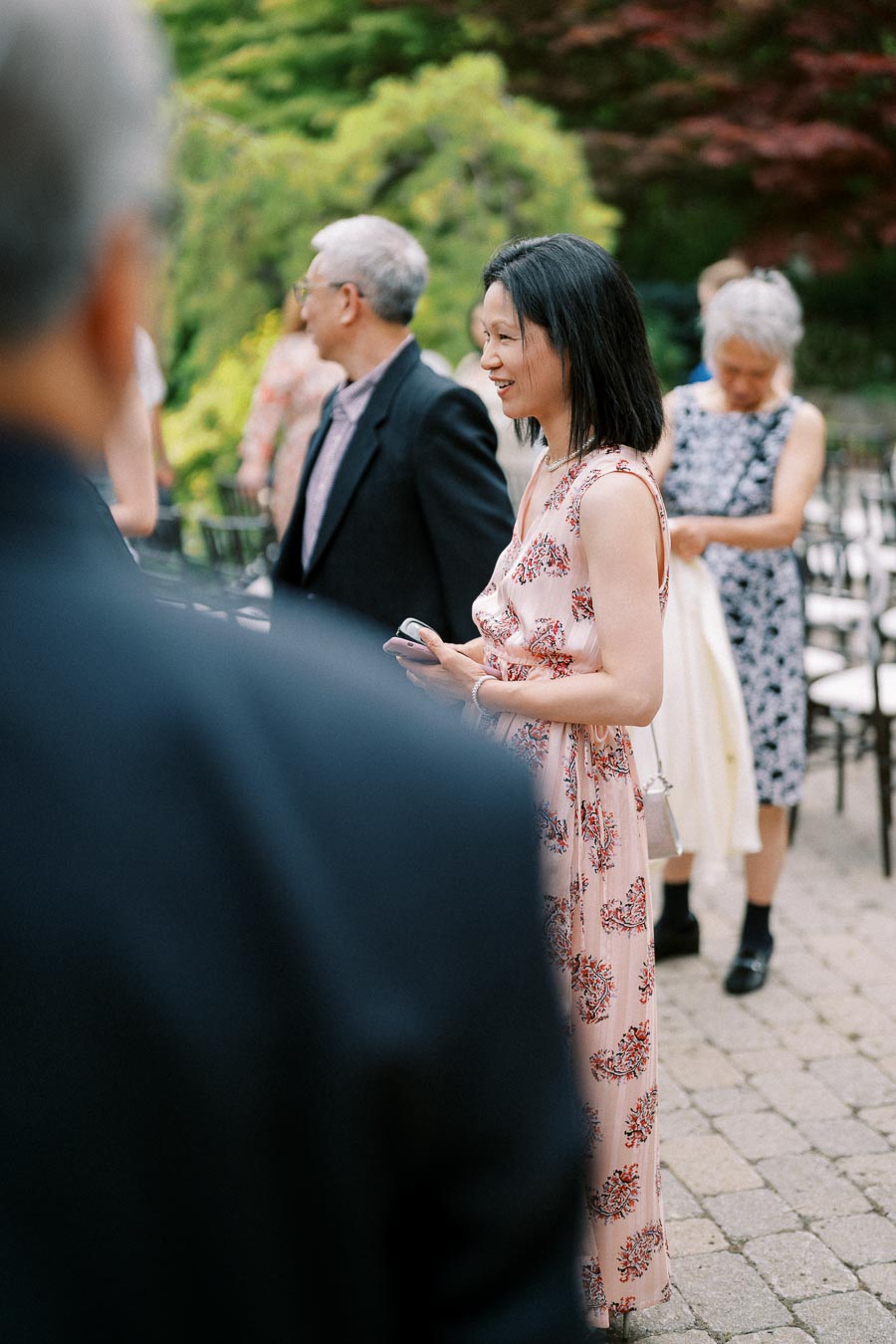 A woman in a floral dress holds her phone while smiling at a garden event, surrounded by other casually dressed guests.