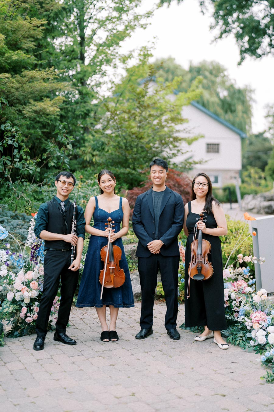 A string quartet posing outdoors, surrounded by lush greenery and colorful flowers, two members holding violins and one holding a clarinet, dressed in formal attire.
