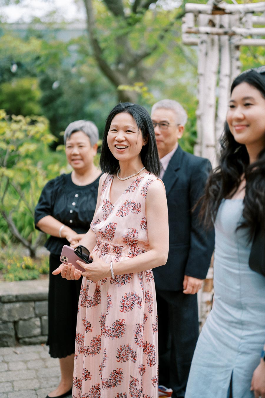 A group of people enjoying an outdoor event, with a woman in a floral dress smiling and holding a phone, surrounded by greenery and a garden setting.