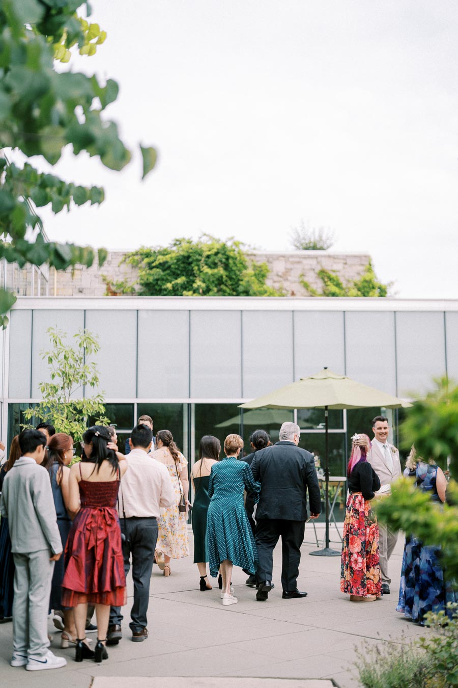 A group of people in formal attire gather outdoors near a modern building with greenery in the background, suggesting a celebration or social event.