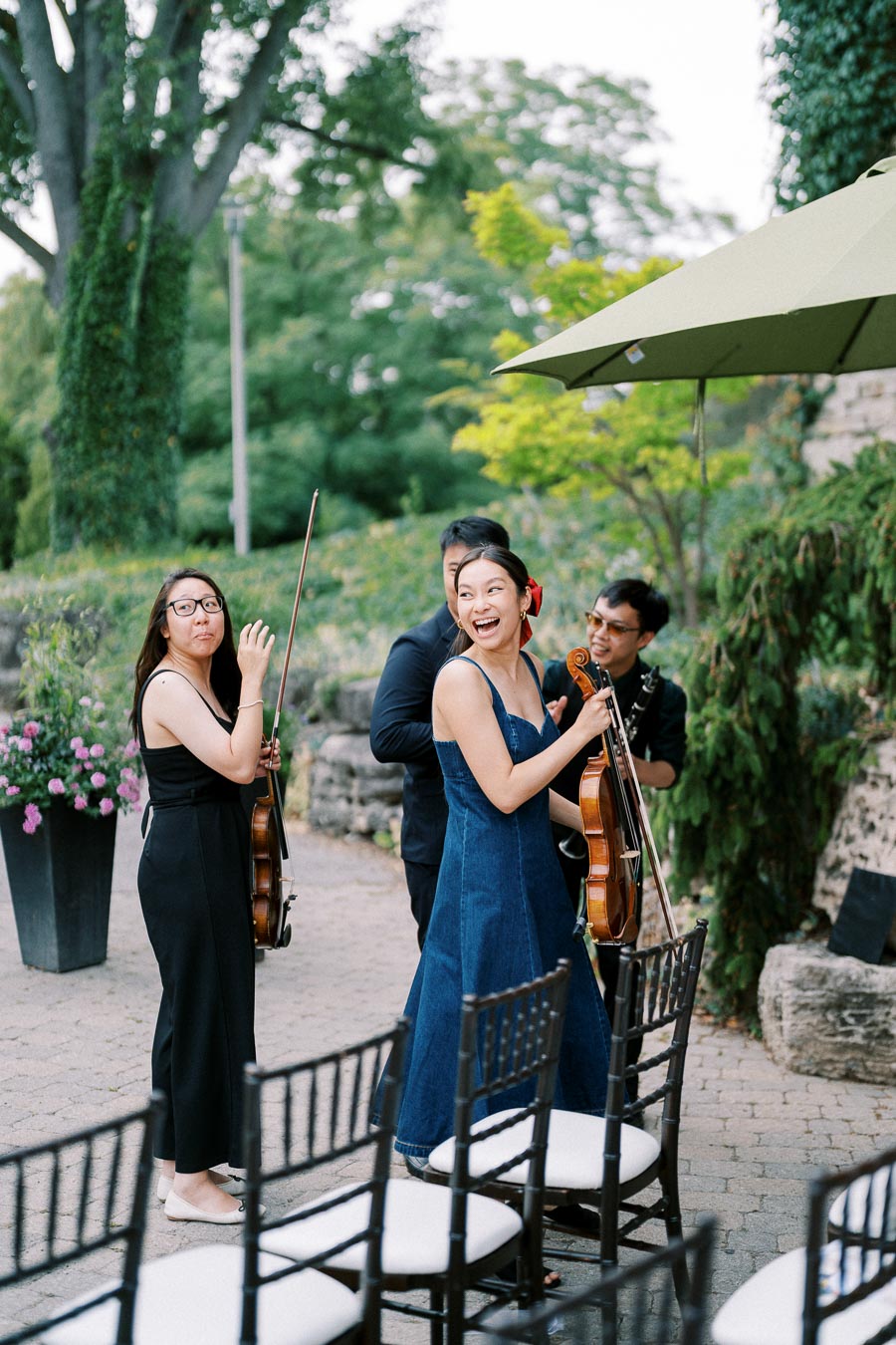 A cheerful string quartet preparing to perform outdoors, with musicians holding violins and a viola, surrounded by lush greenery and elegant seating.