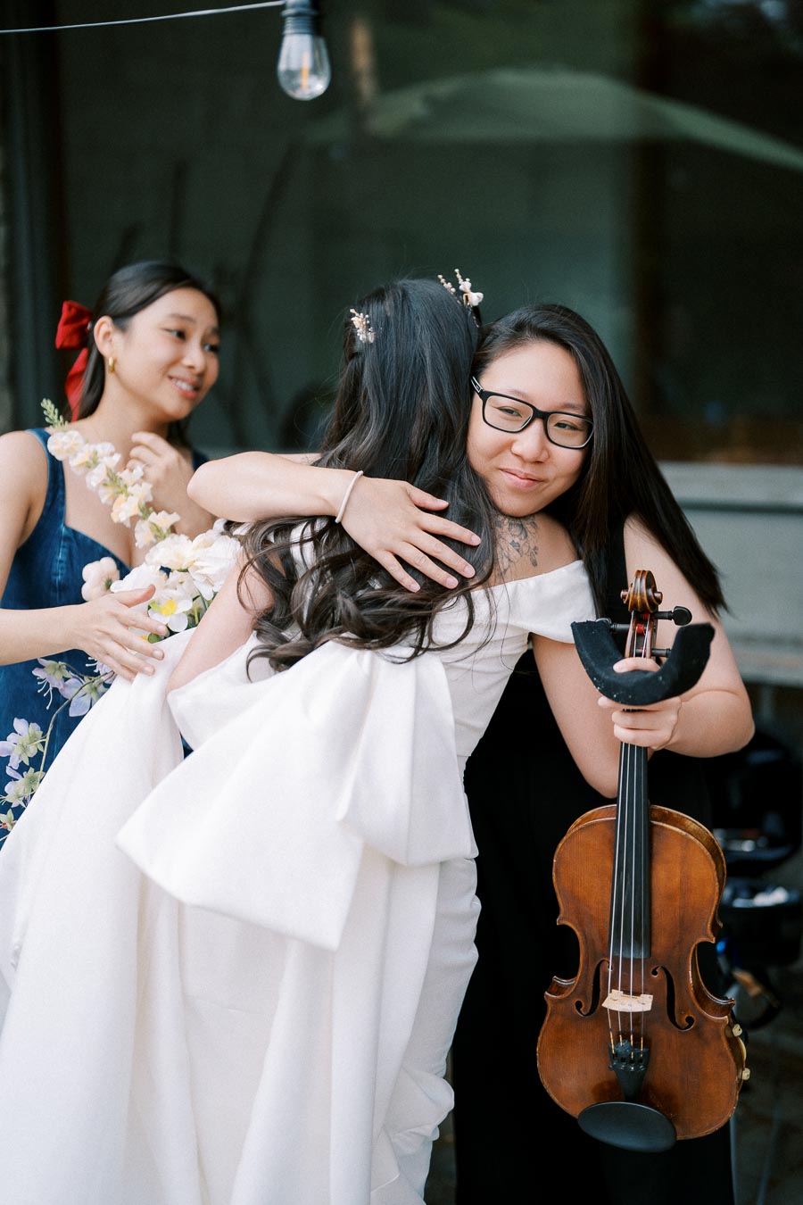 A bride in a white gown embraces a violinist in a heartfelt hug, with a bridesmaid holding a bouquet of flowers nearby, outdoor wedding celebration.