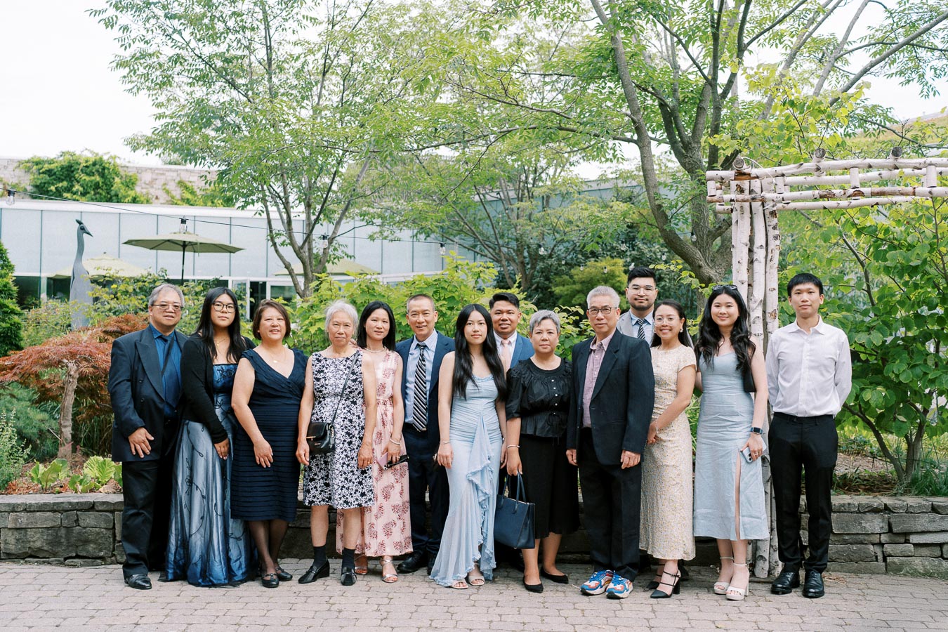 A group of people dressed in formal attire posing for a group photo outdoors in a garden setting with trees and greenery in the background.