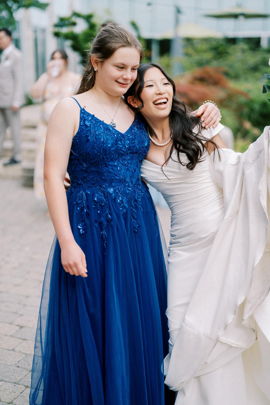 Two friends joyfully embrace, one in a white wedding dress and the other in a blue gown, celebrating together outdoors.