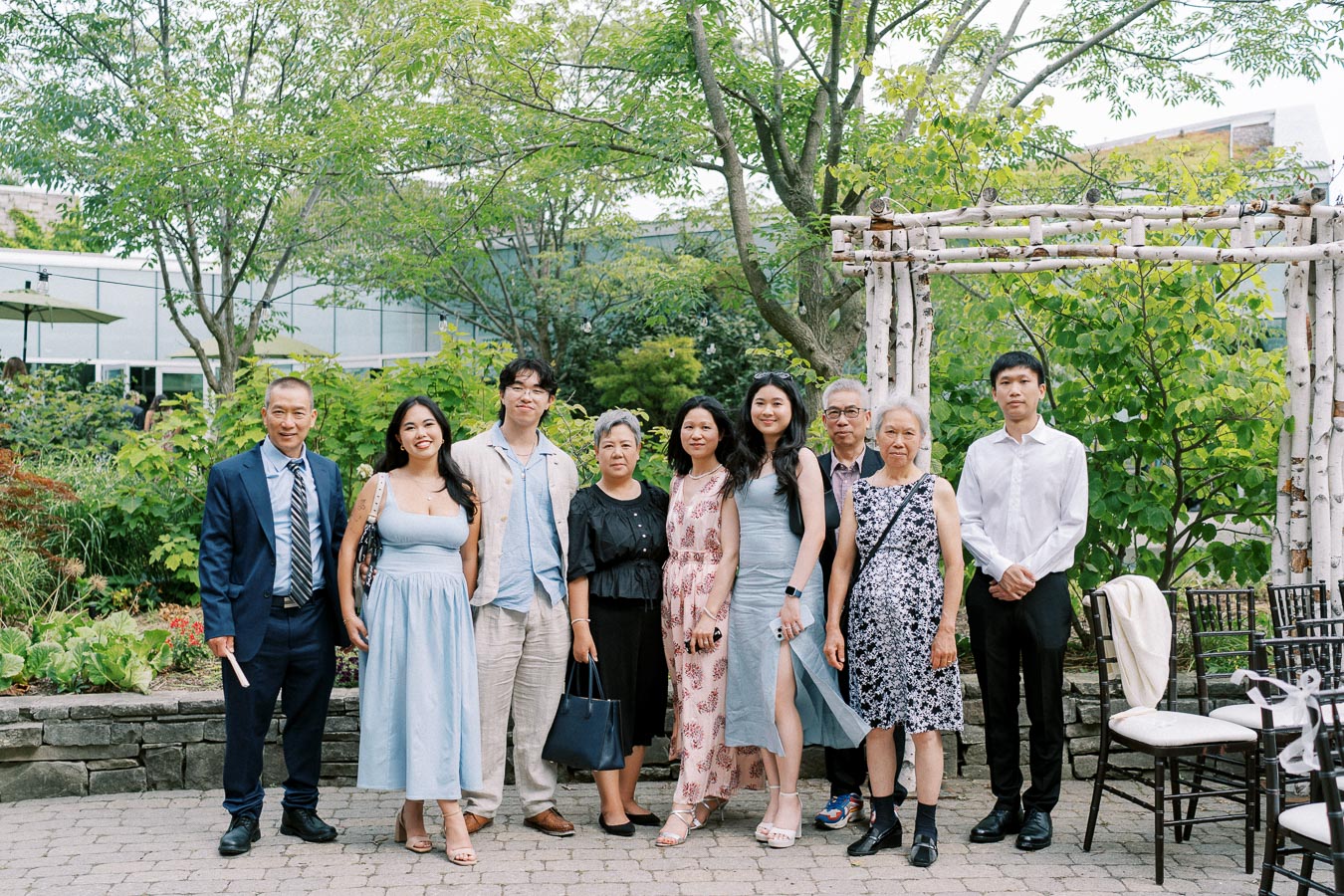 A group of nine people dressed in formal and semi-formal attire, standing together in an outdoor garden setting with lush greenery and a wooden arbor in the background.
