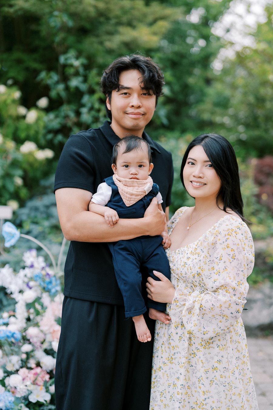 A family portrait featuring a man, woman, and baby standing together outdoors in a lush green garden with colorful flowers in the background. The man is holding the baby, and the woman is smiling, wearing a floral dress.
