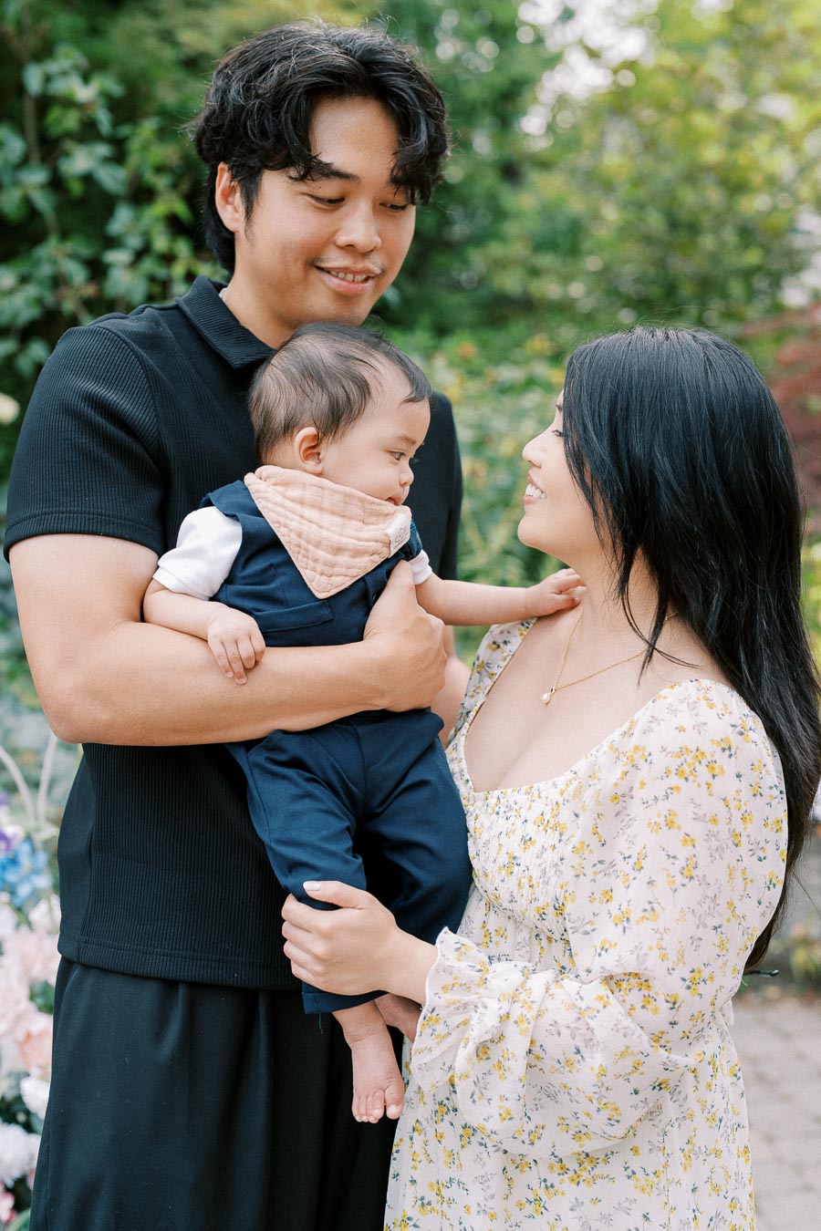 Smiling couple lovingly holding their baby in a garden setting, surrounded by greenery, wearing casual summer attire.