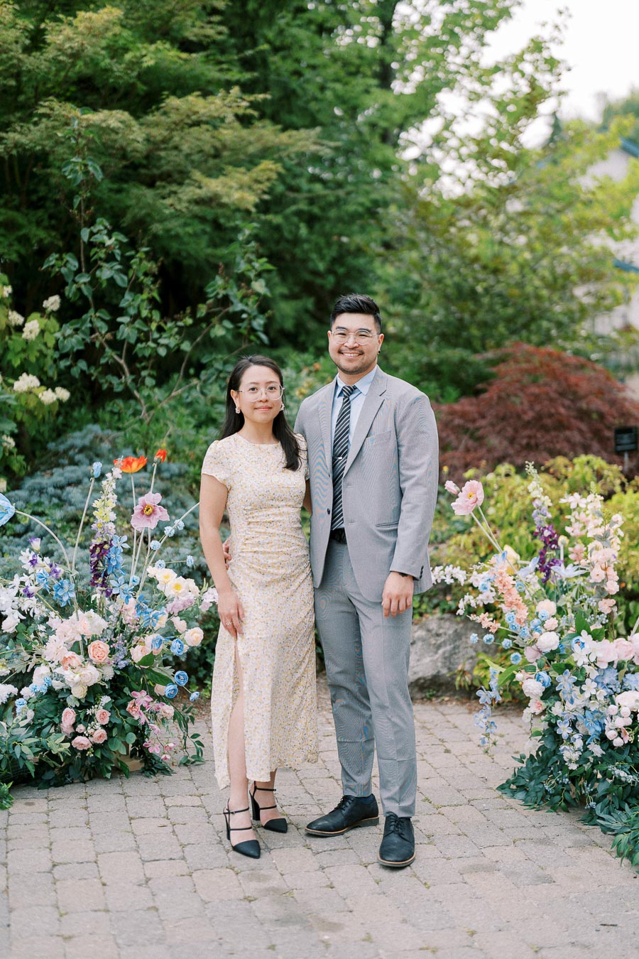 A couple poses happily in a lush garden, surrounded by vibrant flowers and greenery. The woman wears a light floral dress, and the man is in a light gray suit and striped tie. They stand on a stone path with colorful floral arrangements on either side.