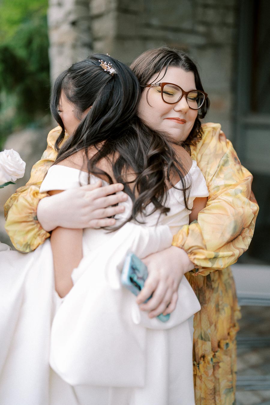 Two women embracing warmly, one wearing a white dress and the other in a yellow floral outfit, standing in front of a stone building. The woman in the white dress is holding a smartphone, and there's a white rose visible in the foreground.