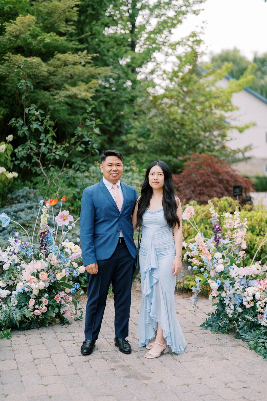 A couple posing outdoors in formal attire, surrounded by vibrant floral arrangements and lush greenery.
