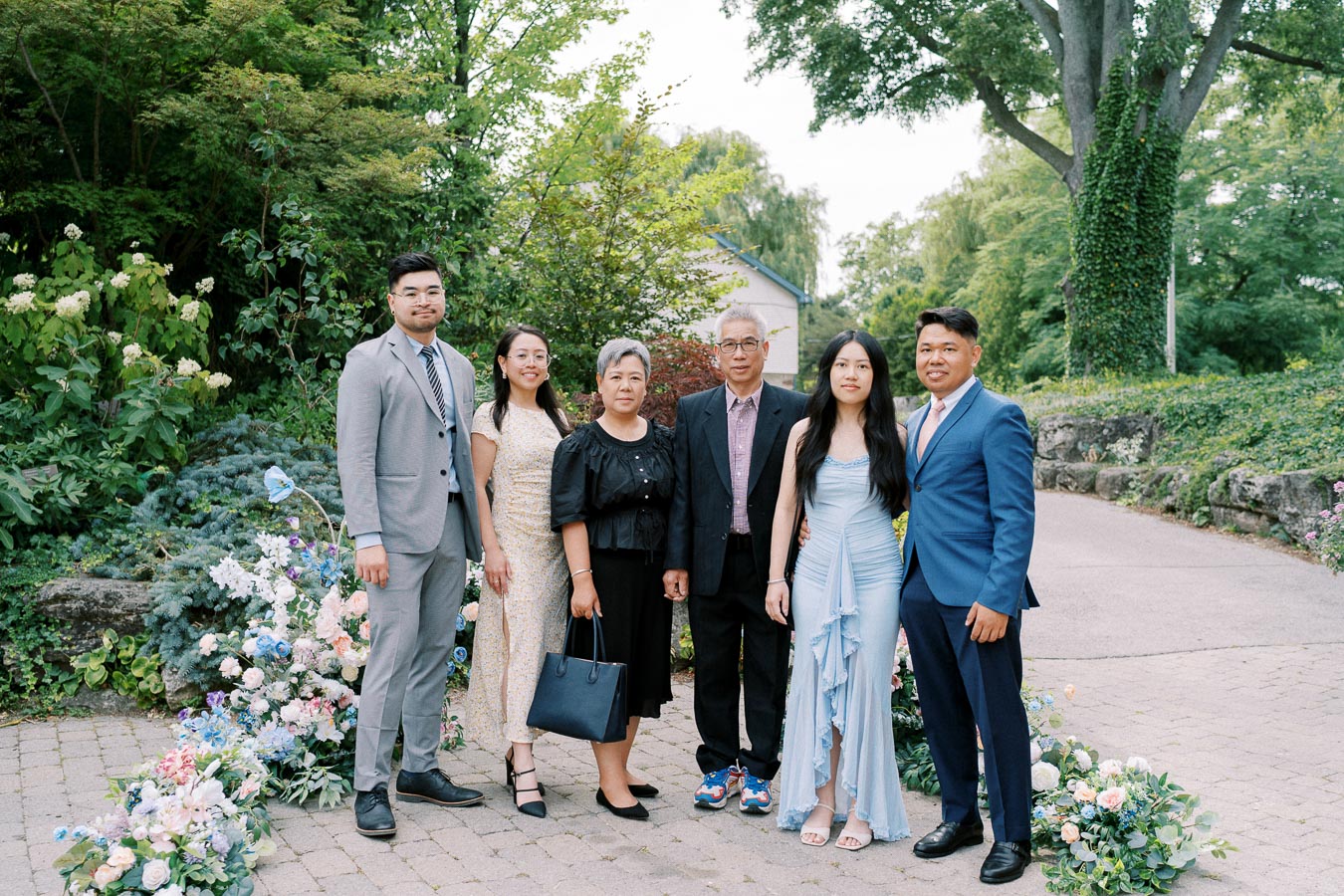 A group of six people, dressed in formal attire, pose together in a lush garden setting with colorful flowers, trees, and a paved pathway.