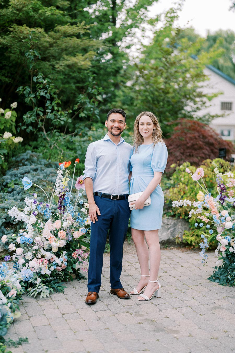 A smiling couple standing on a stone path surrounded by lush greenery and colorful flowers, with the man in a light blue shirt and dark trousers and the woman in a light blue dress holding a clutch.