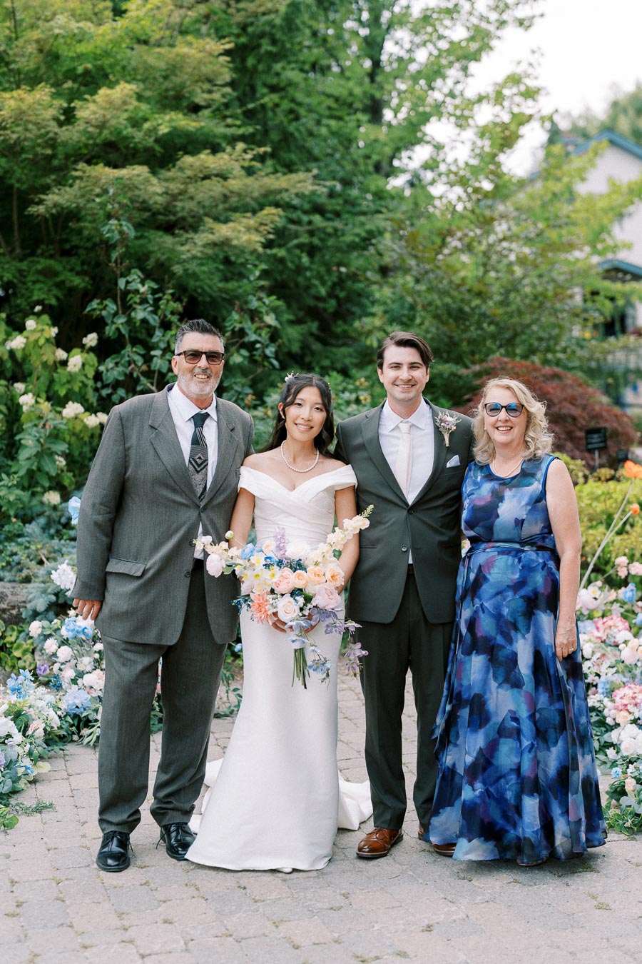 A wedding couple poses with parents in a lush garden setting, featuring blooming flowers and greenery. The bride holds a bouquet of pastel-colored flowers, and the groom wears a dark suit. Both parents are dressed in formal attire, creating a joyful family portrait.