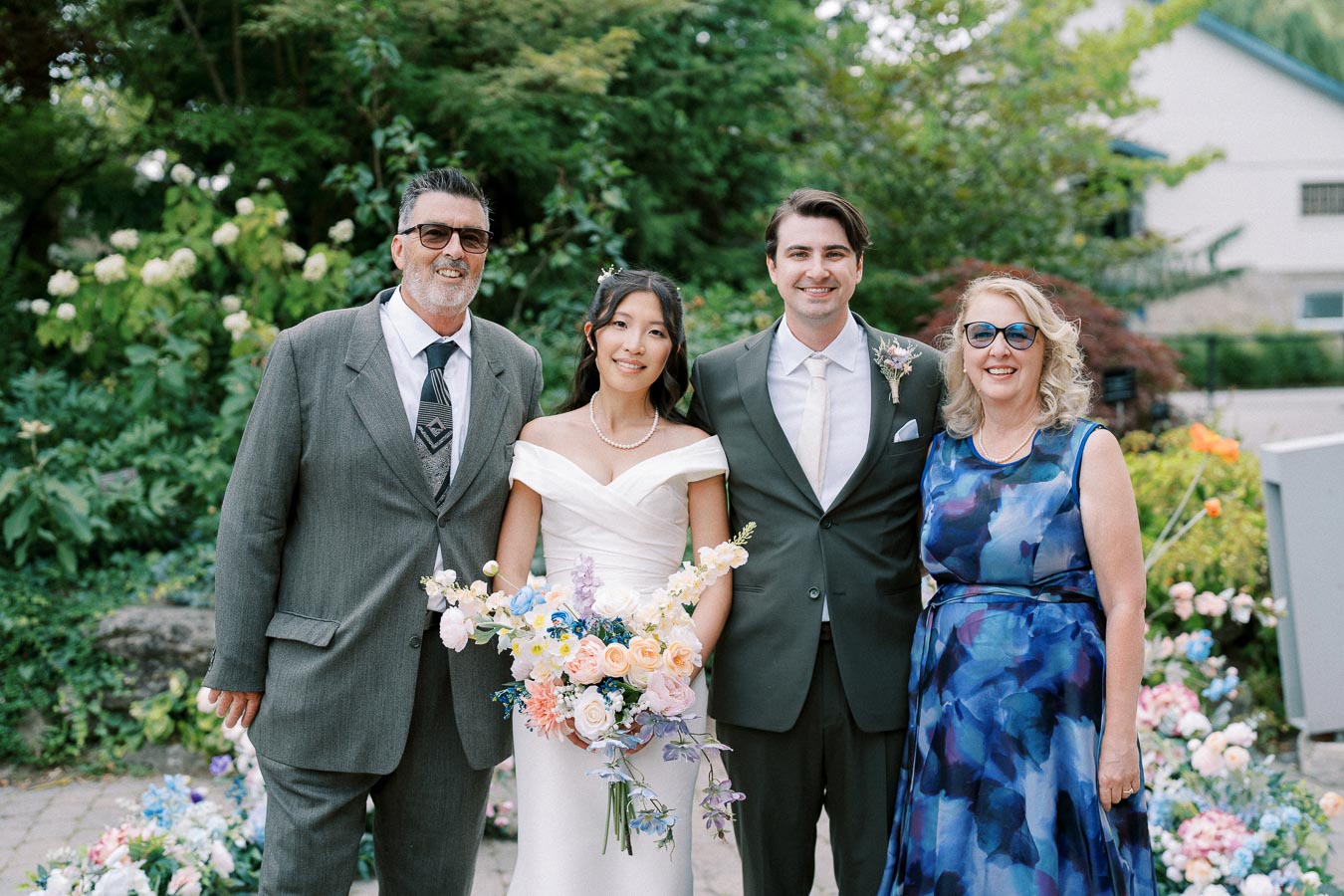 Wedding photo featuring a couple surrounded by family in an outdoor garden setting. The bride holds a bouquet of pastel-colored flowers, and everyone is elegantly dressed, smiling amidst lush greenery and floral decorations.