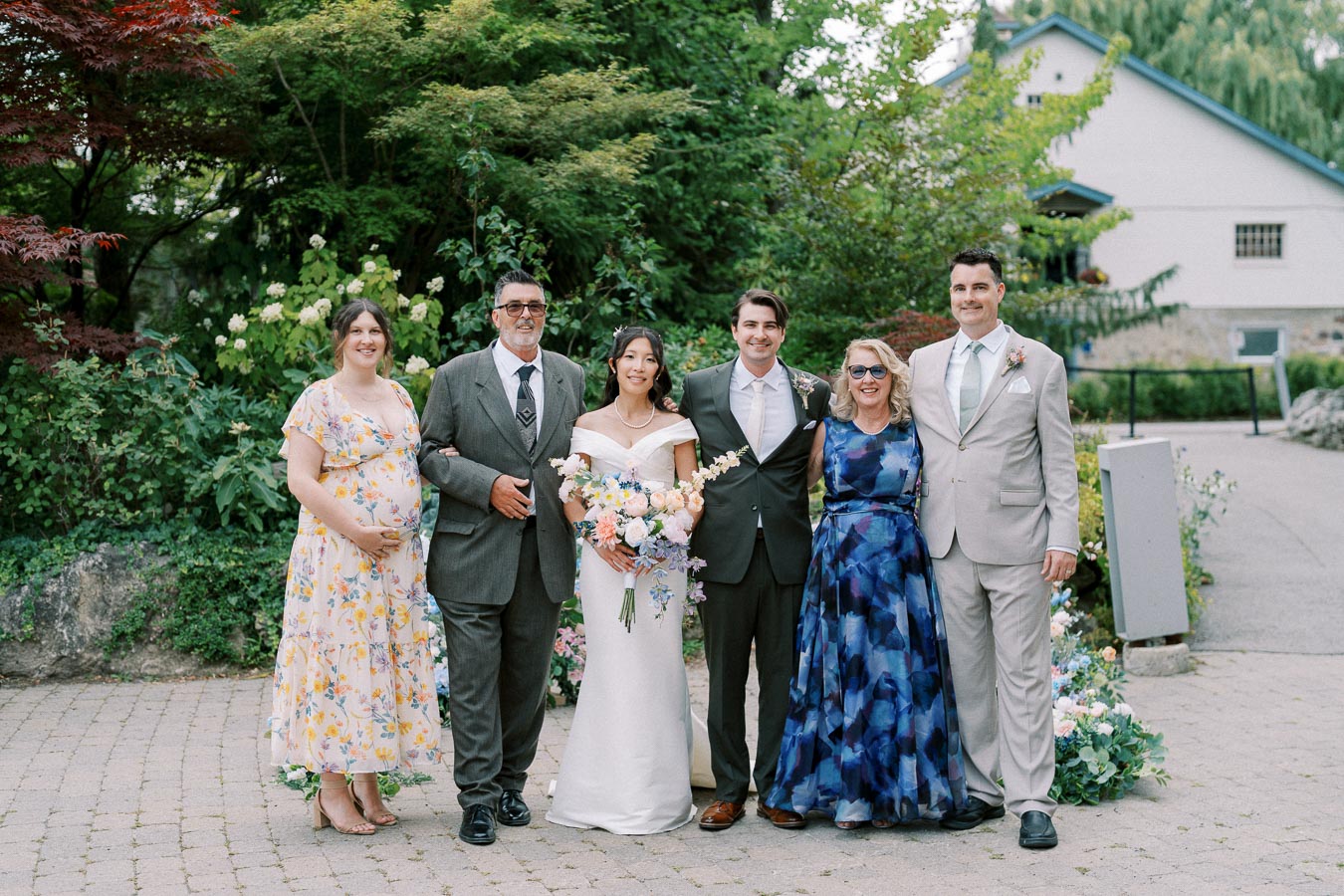 A group portrait of six people, including a bride in a white dress holding a bouquet, a groom in a dark suit, and four other guests in formal attire, posing outdoors in a lush garden setting.