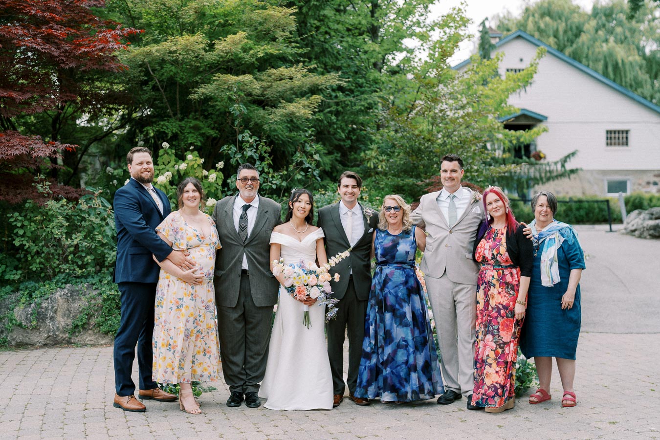 A group photo at an outdoor wedding featuring a bride in a white gown holding a bouquet, standing alongside the groom in a suit. They are surrounded by family and friends dressed in formal wear, set against a lush garden backdrop.