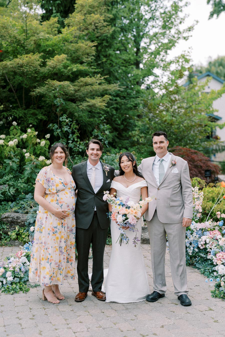 A group of four people stands outdoors, dressed in formal attire, with a lush garden background and colorful flowers. The scene suggests a joyful celebration like a wedding, with one person in a white gown holding a bouquet, and others in suits and a floral dress.