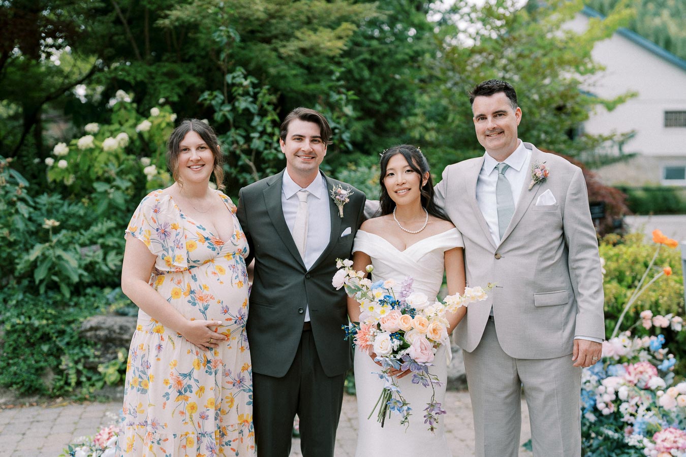 A happy wedding group photo featuring a bride and groom with two friends in a garden setting. The bride holds a colorful bouquet and wears an elegant white dress, while the groom is in a suit and tie. The group is surrounded by lush greenery and vibrant flowers, creating a picturesque outdoor wedding scene.