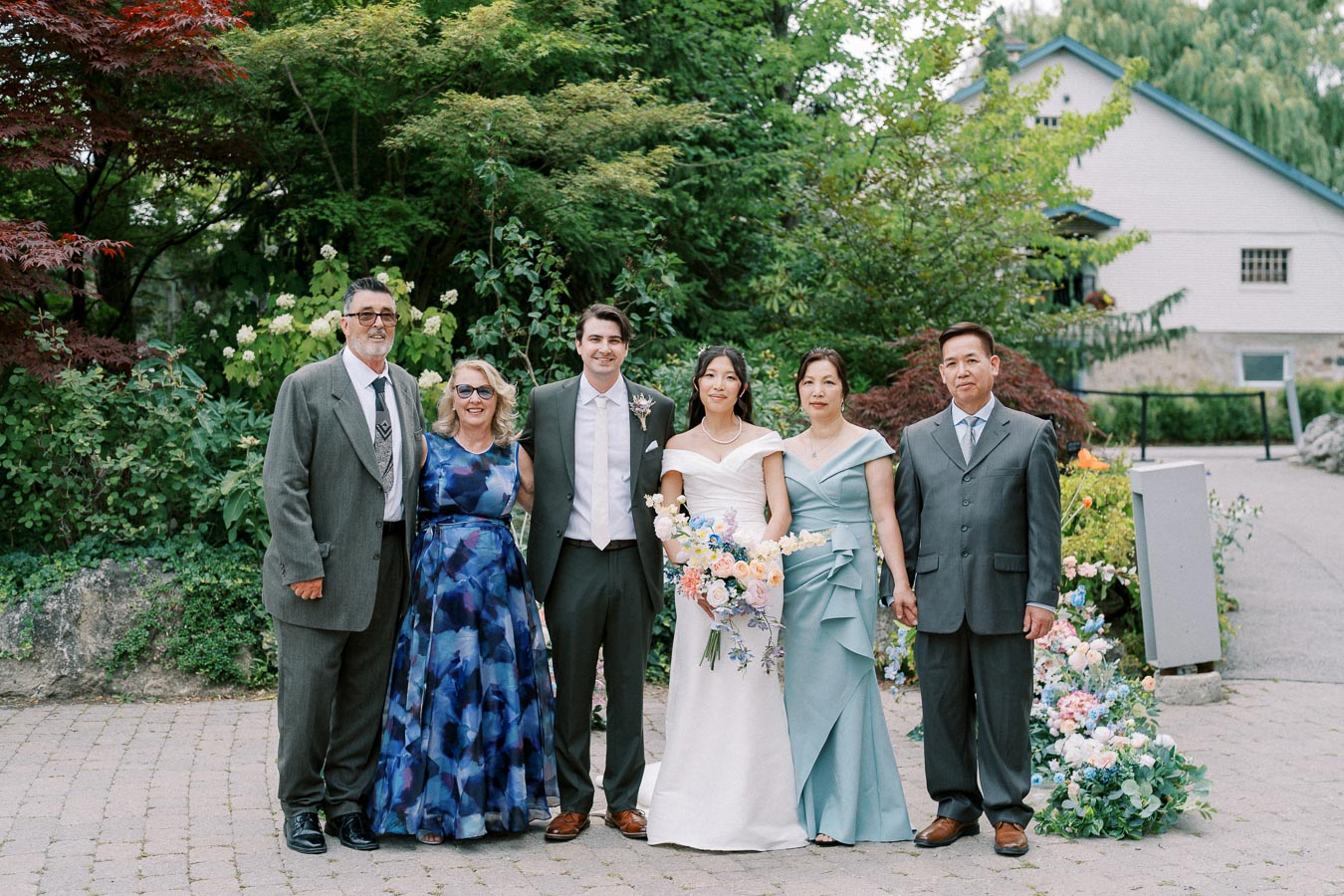Wedding group photo with bride in white gown holding bouquet, groom in dark suit, surrounded by family members in formal attire, set against a lush garden background.