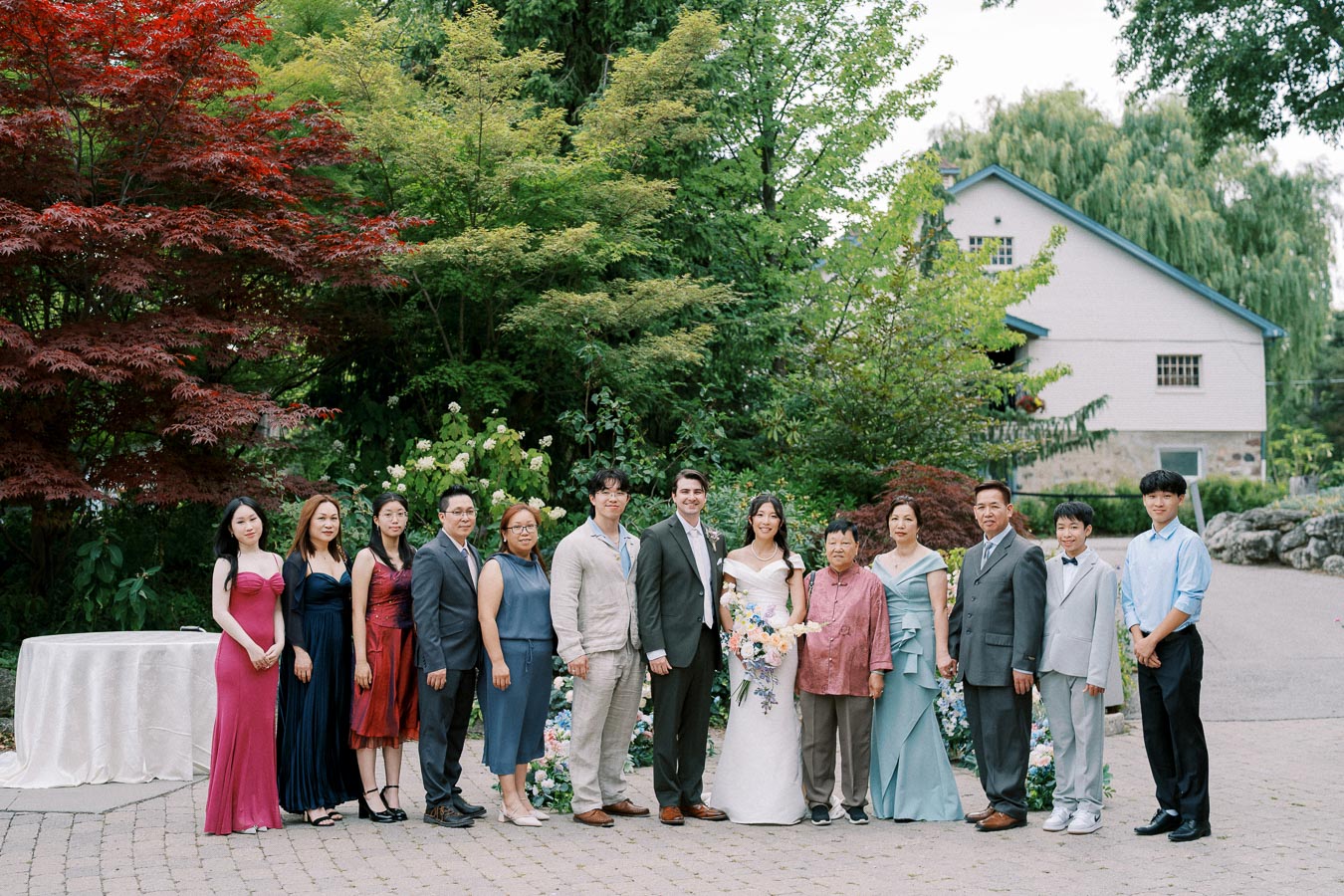 Wedding group photo with the bride and groom surrounded by family and friends, set against a lush garden backdrop featuring vibrant green and red foliage, and a charming house in the background.