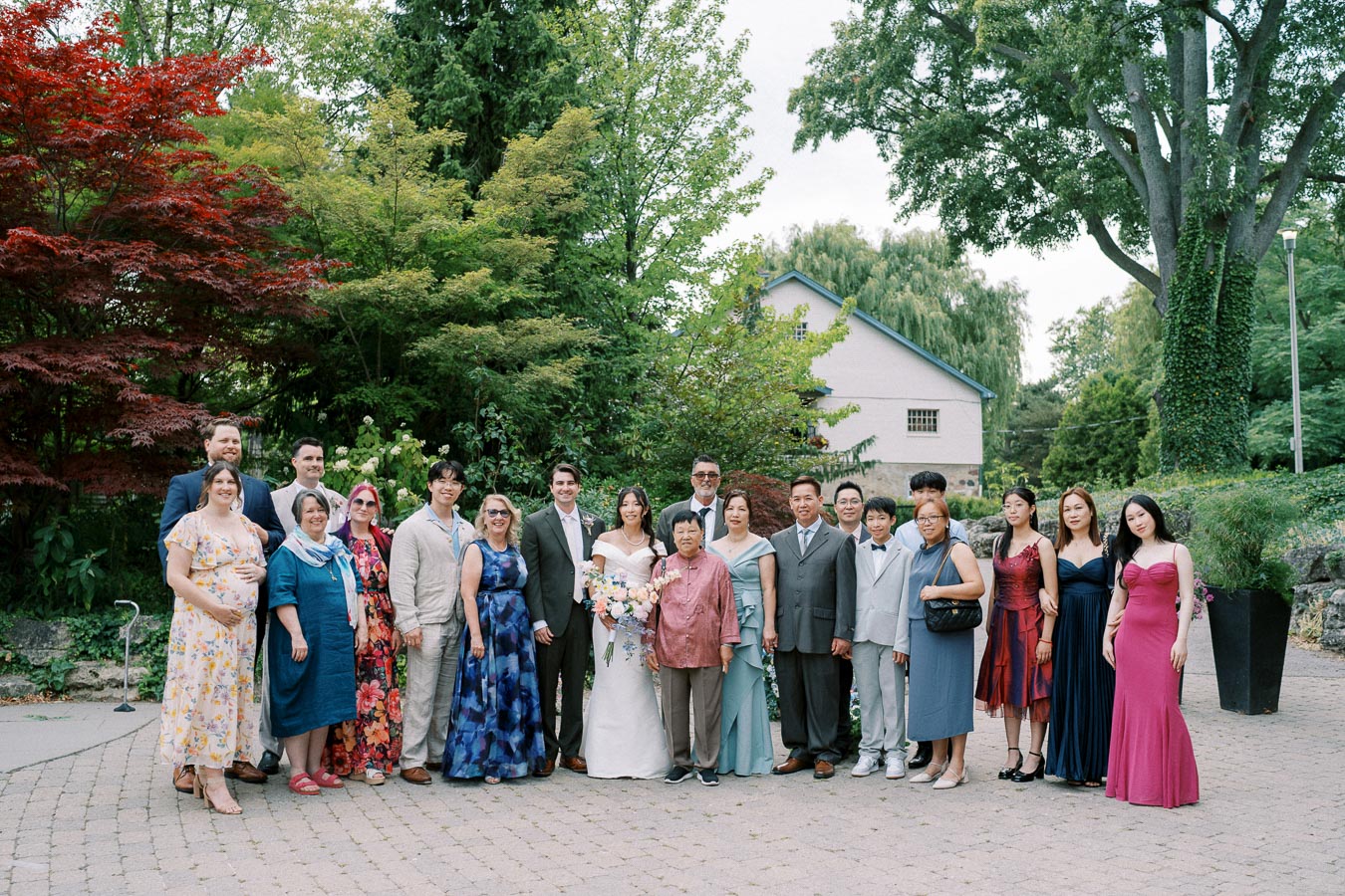 A diverse group of people gathered outdoors for a wedding photo, with the bride and groom in the center surrounded by family and friends, set against a lush garden backdrop.