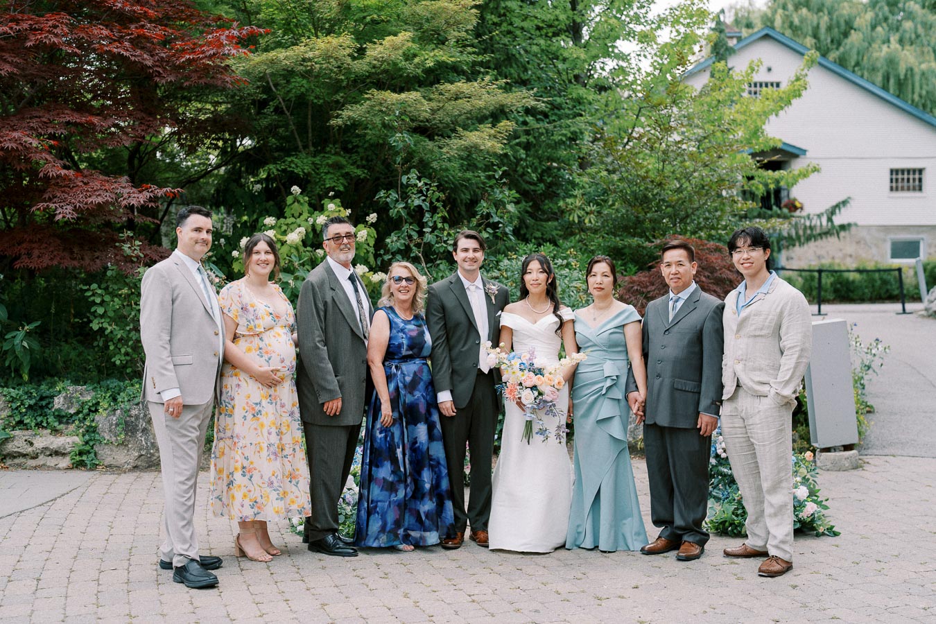 A wedding group photo featuring elegantly dressed guests, including the bride and groom, surrounded by lush greenery and colorful floral arrangements in an outdoor setting.