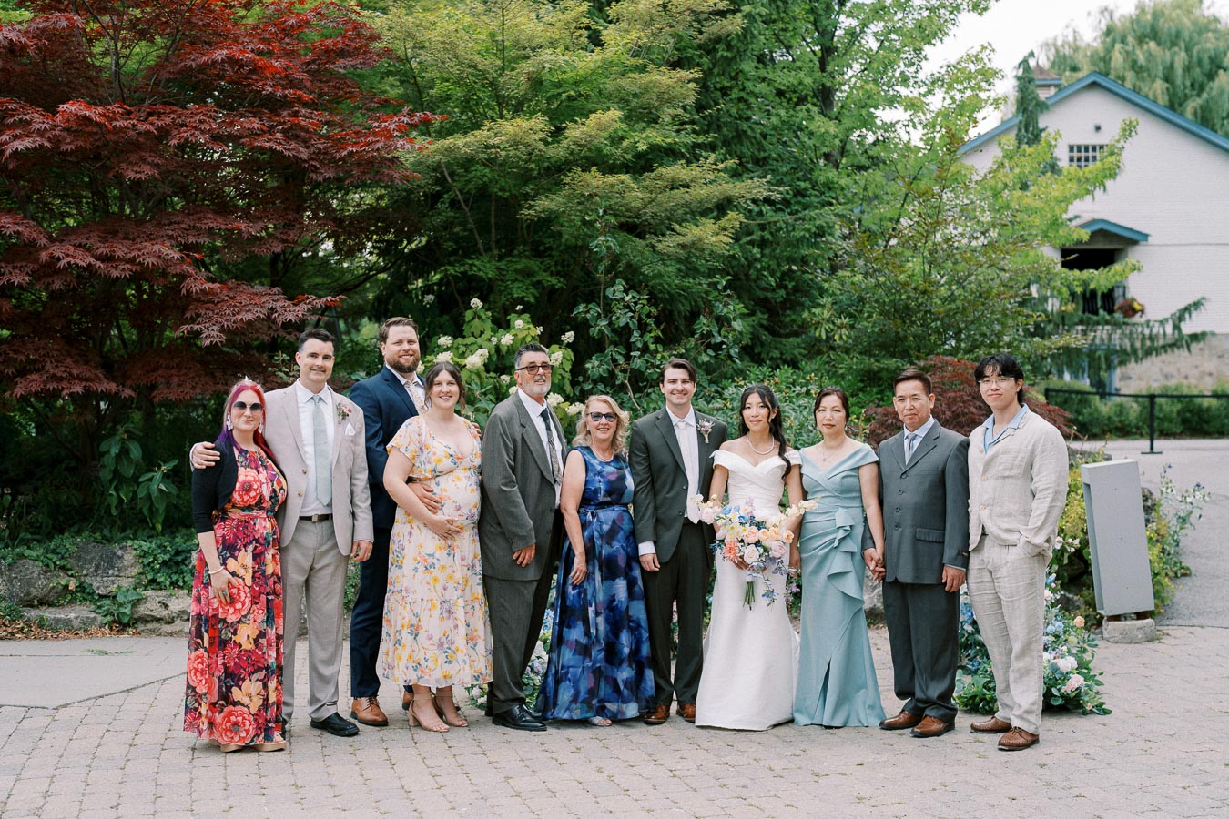 Group photo at an outdoor wedding, featuring guests in formal attire standing together, surrounded by lush green trees and colorful foliage. A bride in a white gown holds a bouquet, accompanied by elegantly dressed family and friends. The setting includes a scenic garden path and a charming house in the background.