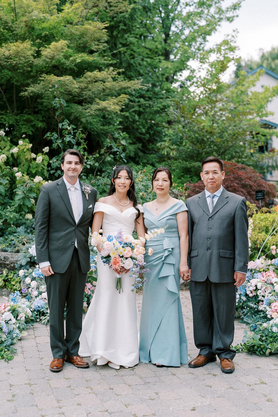 A bride and groom pose with parents in elegant attire amidst a lush garden backdrop. The bride holds a colorful bouquet, and the family stands on a cobblestone path surrounded by greenery and blooming flowers.