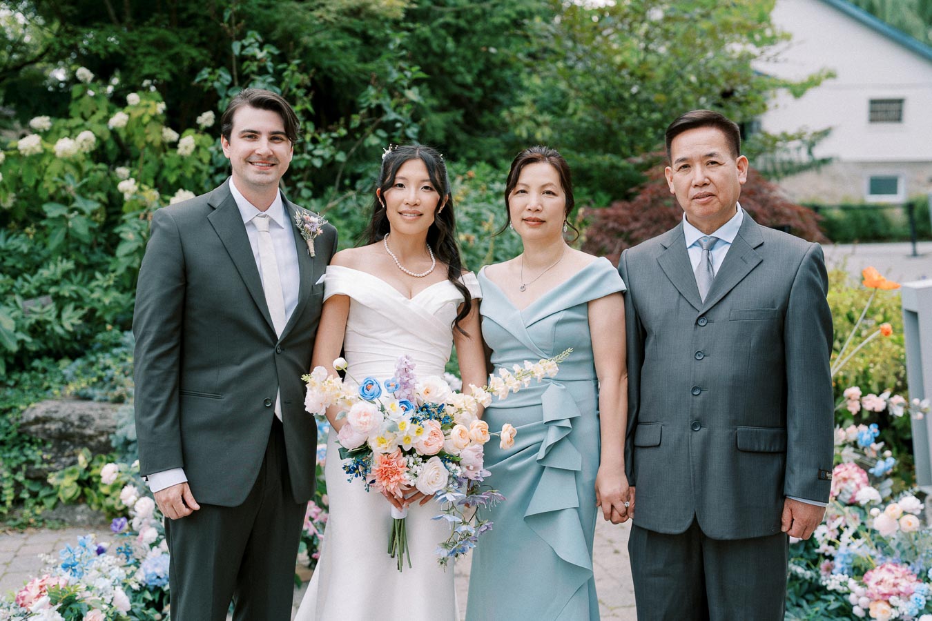 A bride and groom stand outdoors with the bride's parents during a wedding ceremony. The group is surrounded by lush greenery and colorful flowers, with the bride holding a vibrant bouquet. The groom and father are dressed in suits, while the bride and mother wear elegant gowns.