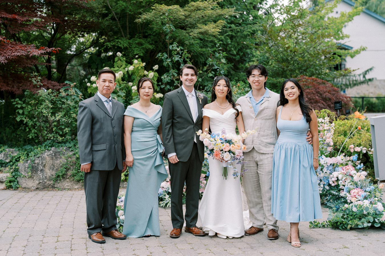 A group photo at an outdoor wedding, featuring six people dressed in formal attire, with a bride in a white gown holding a bouquet of colorful flowers, standing beside a groom in a dark suit. The image is set against a lush garden backdrop with vibrant greenery and flower arrangements.