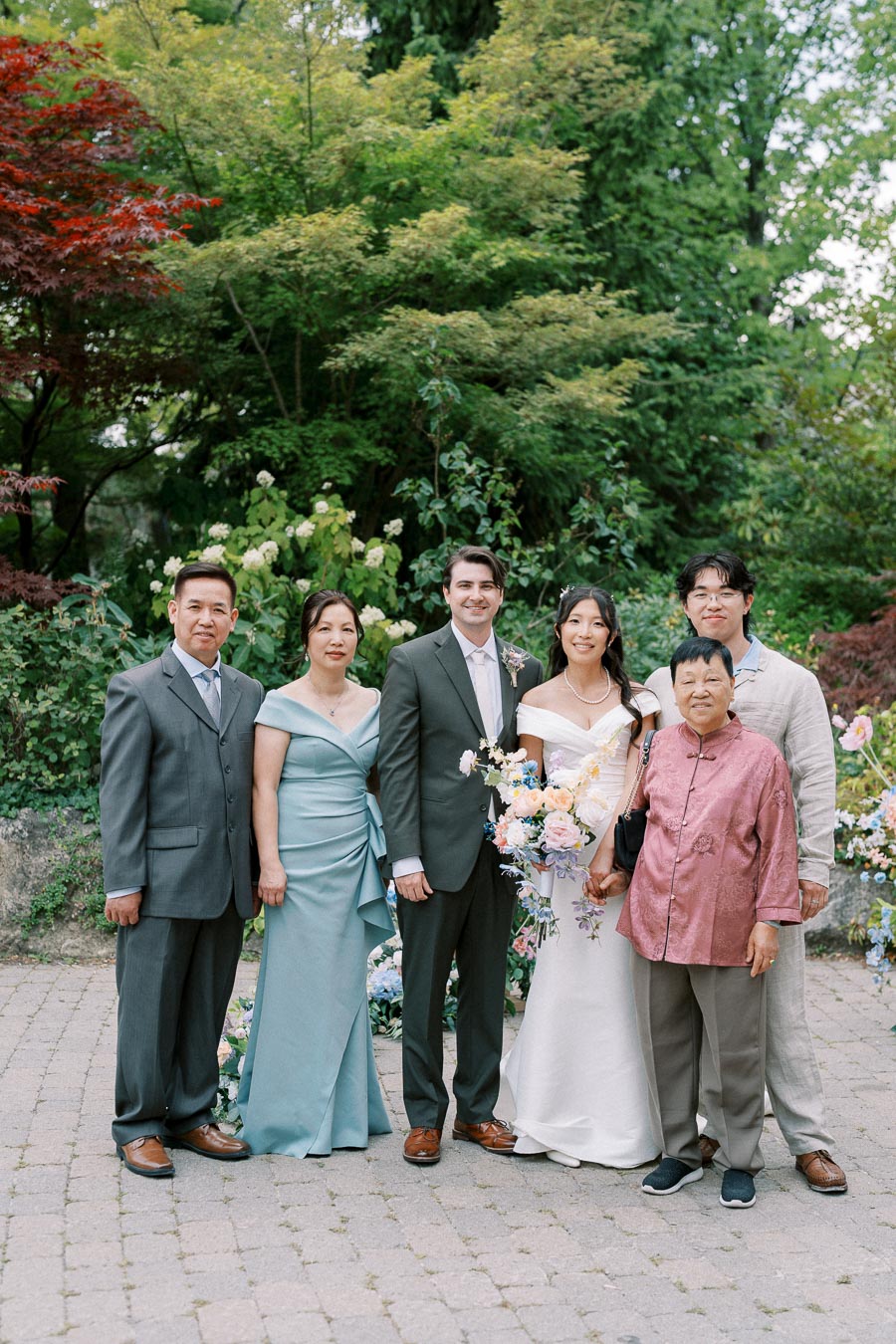 A wedding group photo featuring a bride and groom surrounded by family members in an outdoor garden setting with lush greenery and blooming flowers.