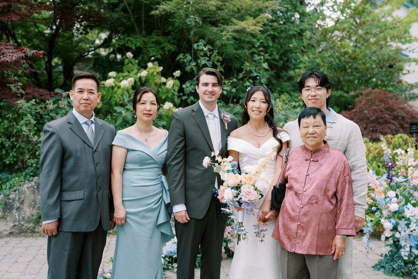 A wedding group photo in a lush garden setting, featuring a bride in a white dress holding a bouquet of pastel flowers, a groom in a suit, and four guests dressed in formal attire, surrounded by vibrant greenery and floral arrangements.