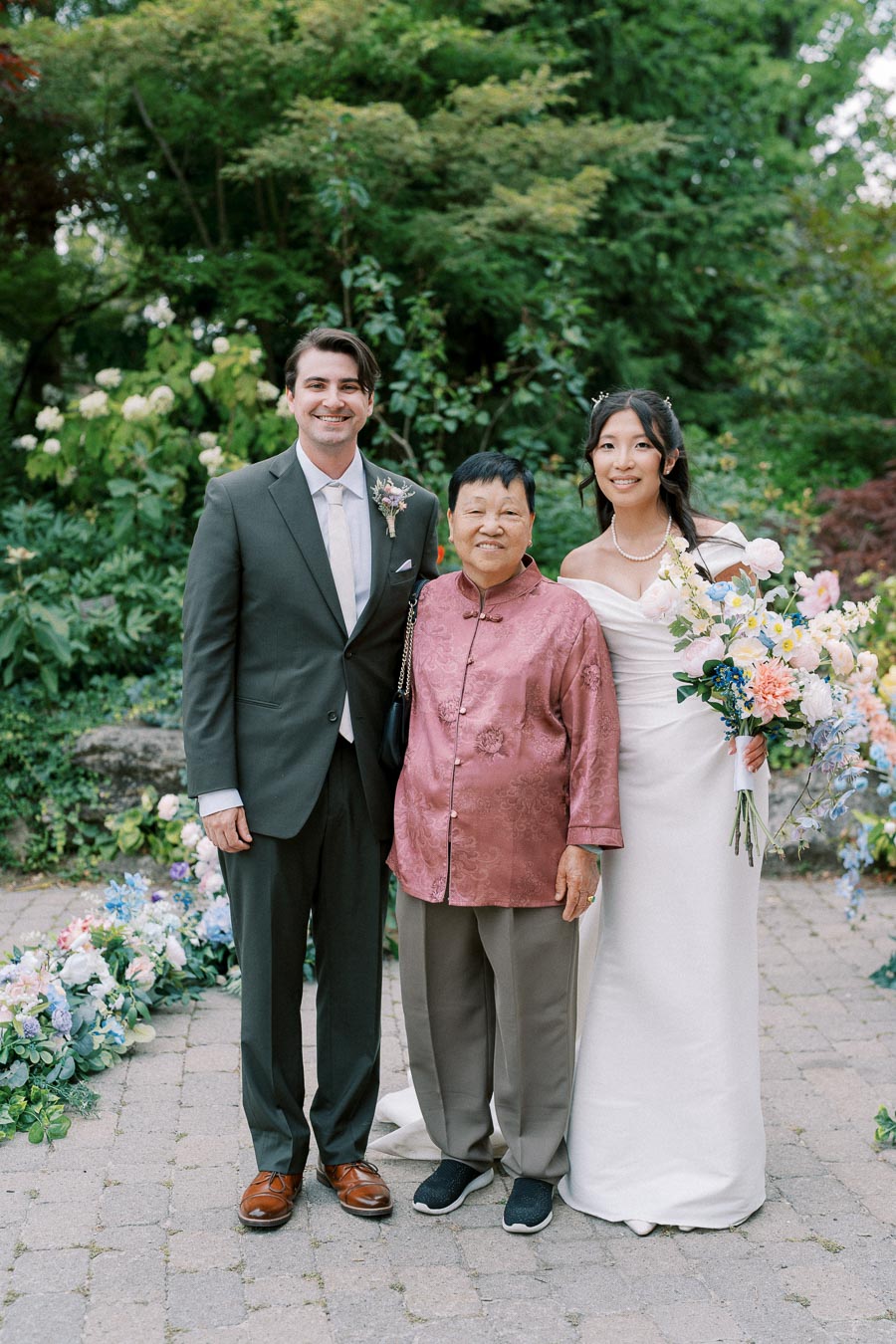 Wedded couple posing with family member in scenic garden, the groom in a dark suit and the bride in an off-shoulder white gown holding a colorful bouquet, surrounded by lush greenery and floral arrangements.