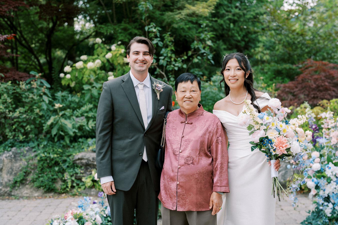 Wedding couple and guest posing in garden setting, bride holding colorful bouquet, showcasing lush greenery and floral arrangements.