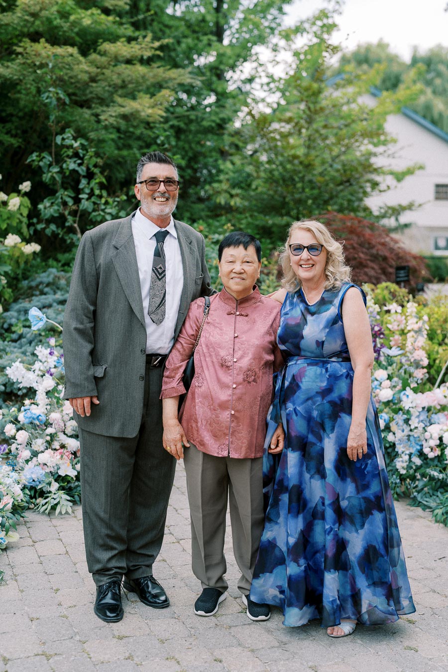 Three people smiling at a garden party, with two women in elegant attire and a man in a suit standing on a stone path surrounded by lush greenery and colorful flowers.