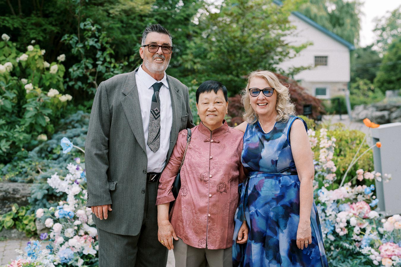 Three people smiling and posing in a garden setting, surrounded by vibrant flowers and greenery, with a building visible in the background. They appear to be enjoying a special occasion or event.