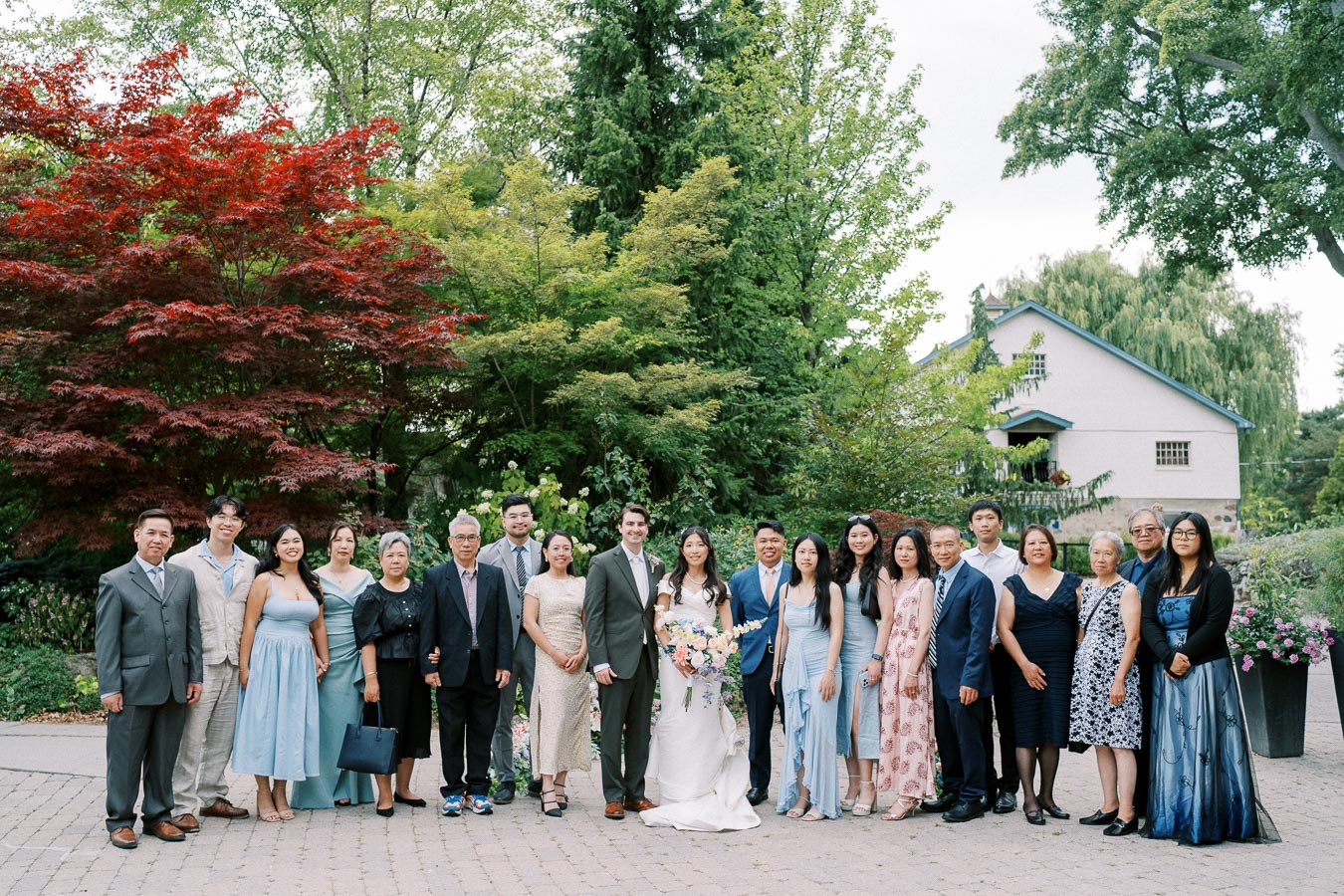 A joyful wedding party group poses outdoors in front of lush green and red foliage, with the bride and groom standing at the center, surrounded by family and friends in elegant attire, capturing a celebratory moment in a garden setting.
