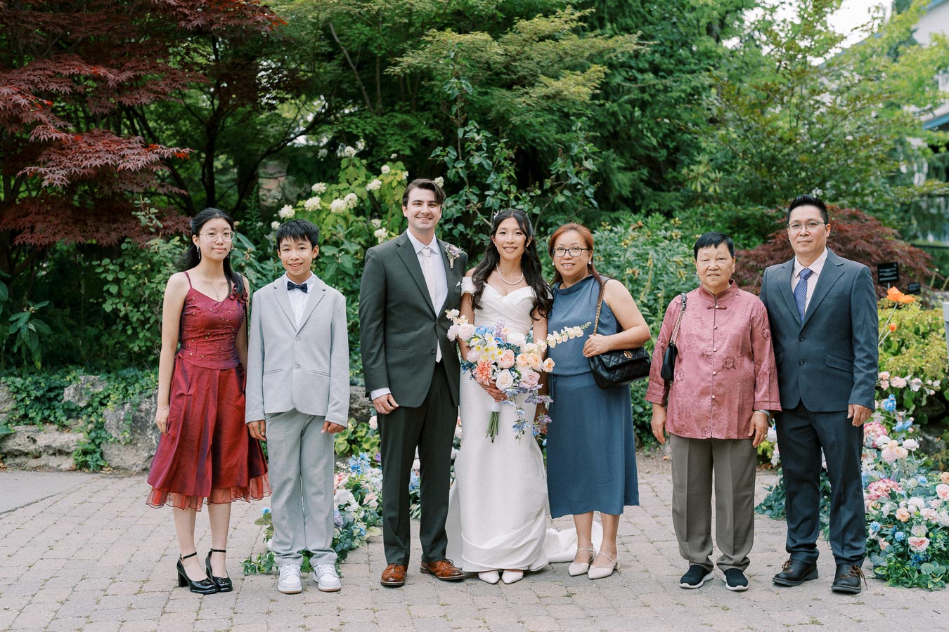 A family gathered together in a lush outdoor garden setting, dressed in formal attire. The bride holds a colorful bouquet, standing alongside the groom and family members. The scene is framed by vibrant greenery and blooming flowers, creating a celebratory atmosphere.