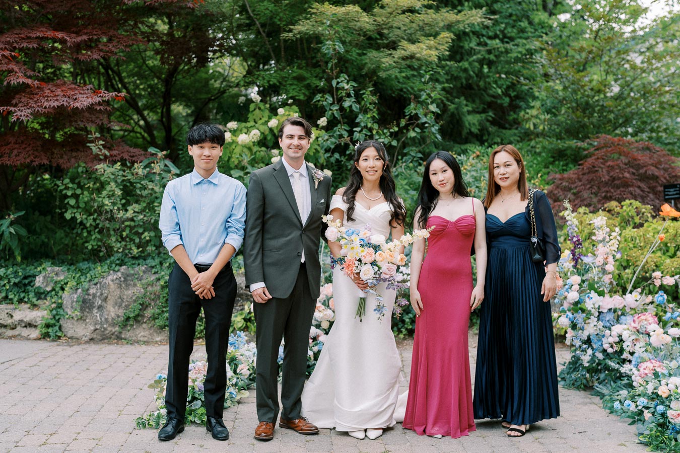A group of five people posing outdoors in formal attire at a wedding ceremony, surrounded by lush greenery and colorful floral arrangements.