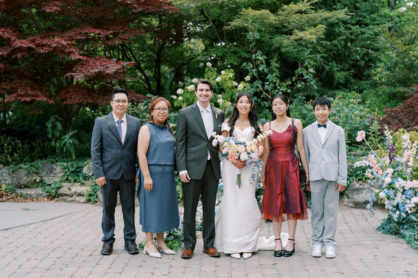 A group photo of people in formal attire at an outdoor wedding, featuring a bride in a white gown holding a bouquet, a groom in a dark suit, and family members standing on a paved garden path surrounded by lush greenery and colorful flowers.