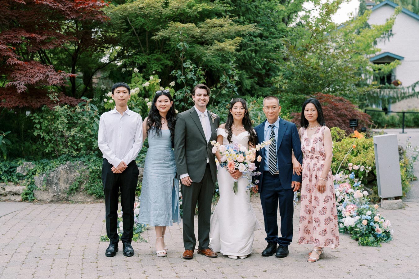 A group photo of a wedding party in a lush garden setting. The bride, wearing a white gown and holding a bouquet of flowers, stands next to the groom in a suit. Flanked by family members, they pose on a cobblestone path surrounded by vibrant greenery and floral arrangements.