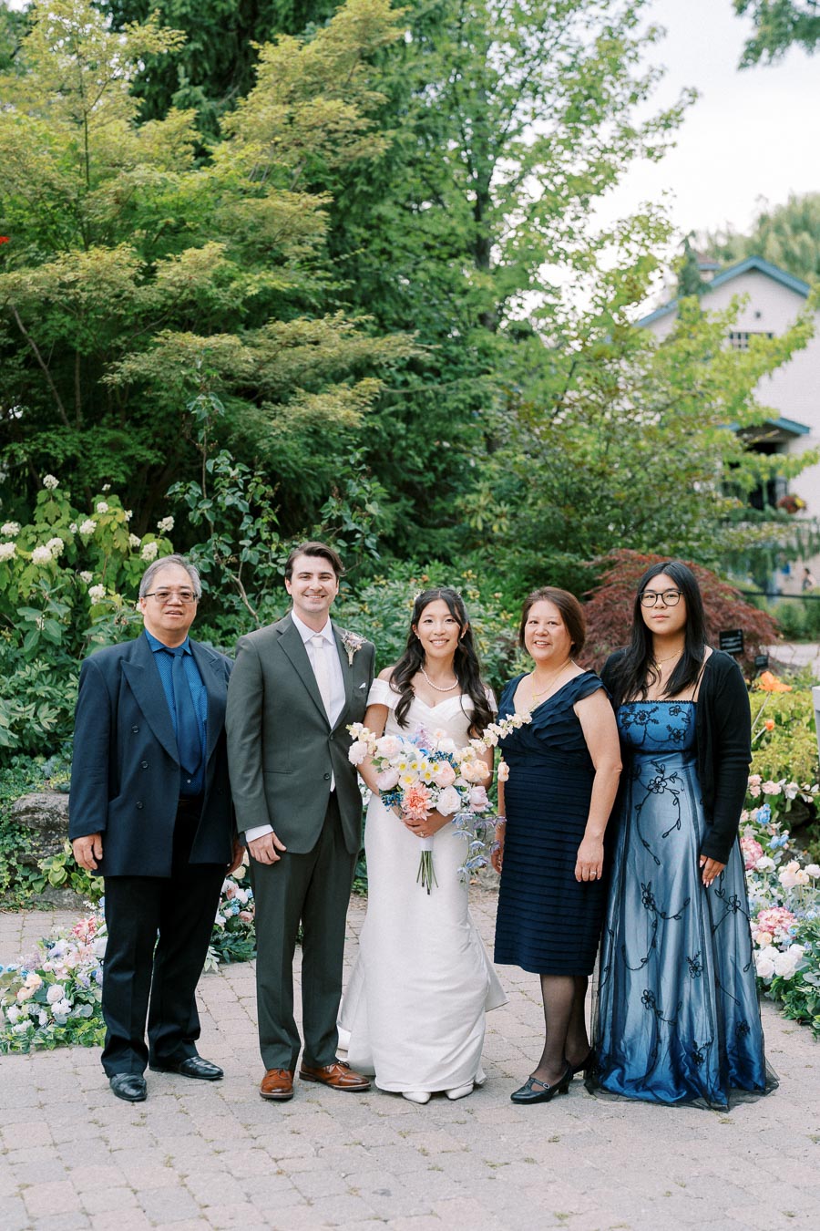 Wedding group photo in a lush garden setting, featuring a bride in a white dress holding a vibrant bouquet, a groom in a gray suit, and three family members elegantly dressed, surrounded by blooming flowers and greenery.