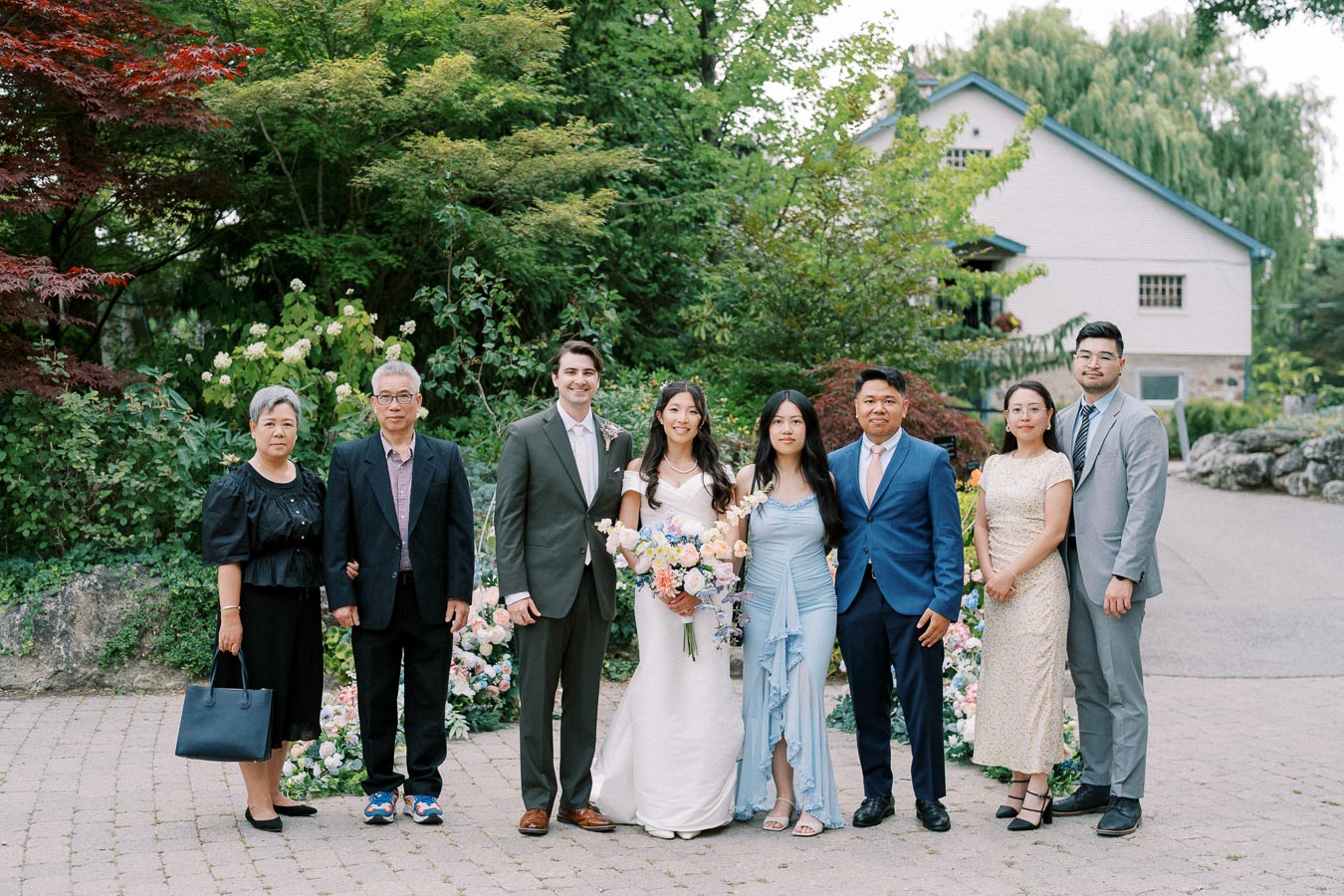 A group of people posing for a formal family photo at an outdoor wedding, with lush greenery and a house in the background. The bride stands in a white gown holding a bouquet, surrounded by smiling family members dressed in suits and elegant dresses.