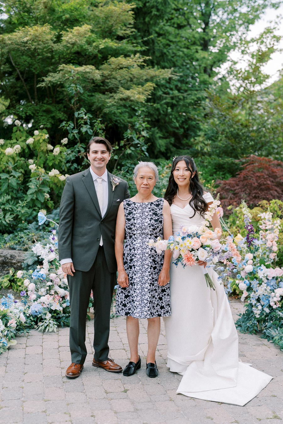 Three people smiling in a garden setting, including a bride in a white dress holding a colorful bouquet, a groom in a dark suit, and an elderly woman in a patterned dress, surrounded by lush greenery and flowers.