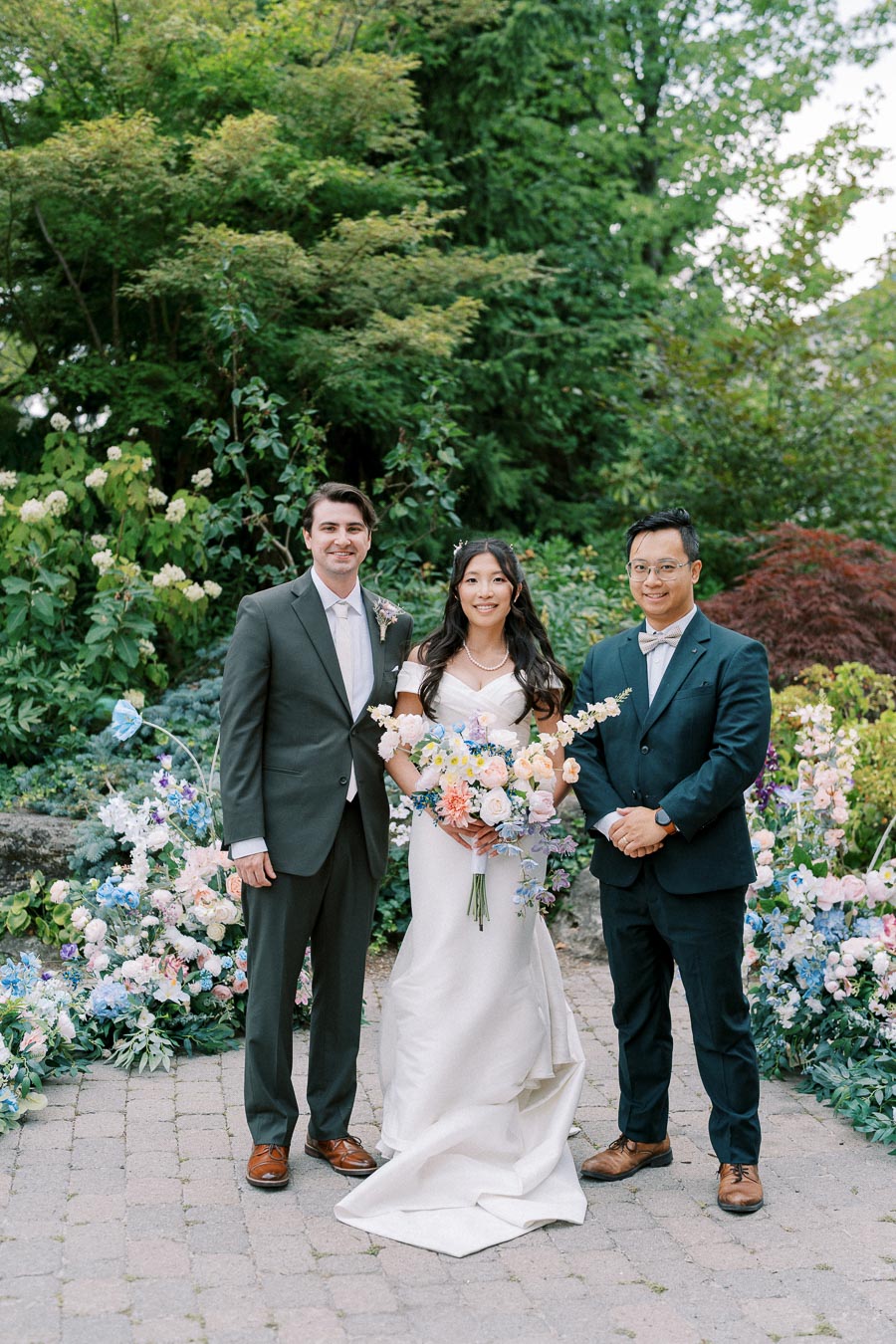 Wedding ceremony in a lush garden setting with a bride in a white gown holding a colorful bouquet, flanked by two groomsmen in suits.