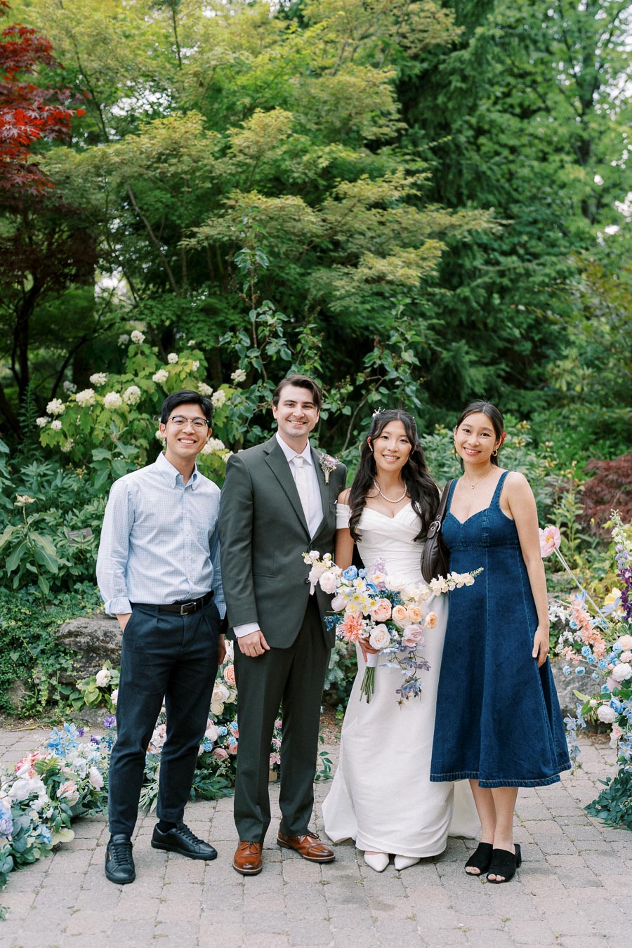 A bride in a white dress and groom in a suit pose with two guests in a lush, green garden decorated with colorful flowers.
