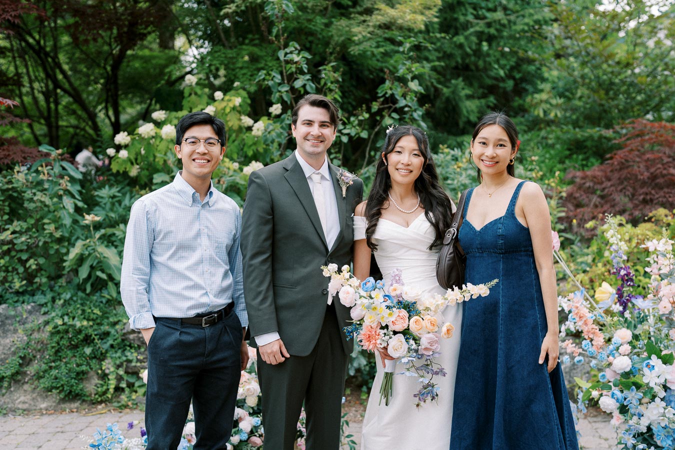 A wedding couple poses with two guests in a lush garden, surrounded by colorful flowers. The bride in a white gown holds a vibrant bouquet, while the groom wears a dark suit. The two guests are dressed in smart casual attire, smiling warmly amidst the scenic backdrop of greenery and blossoms.